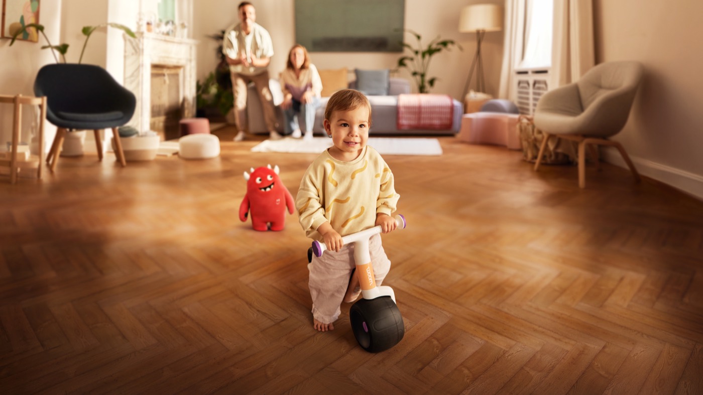 A toddler on a Woom WOW balance bike indoors with family nearby