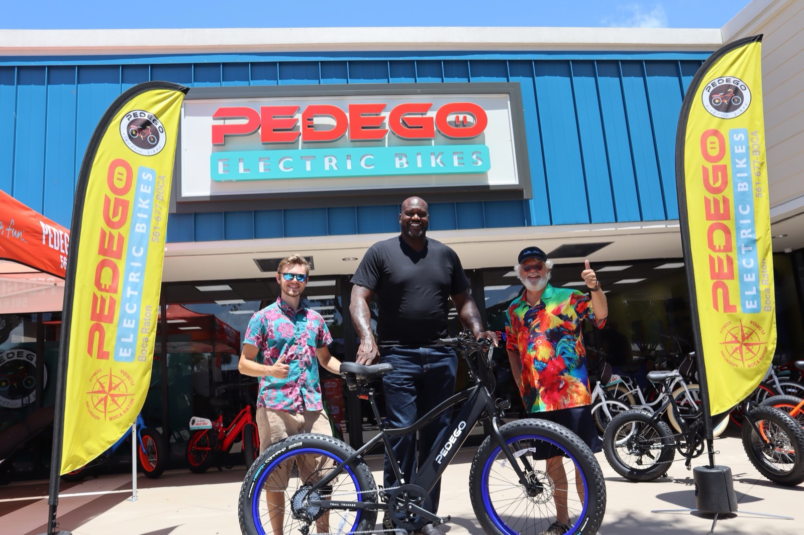 Shaquille O'Neal outside Pedego Boca with Kyle and Tom, standing behind a Pedego Trail Tracker fat-tire e-bike